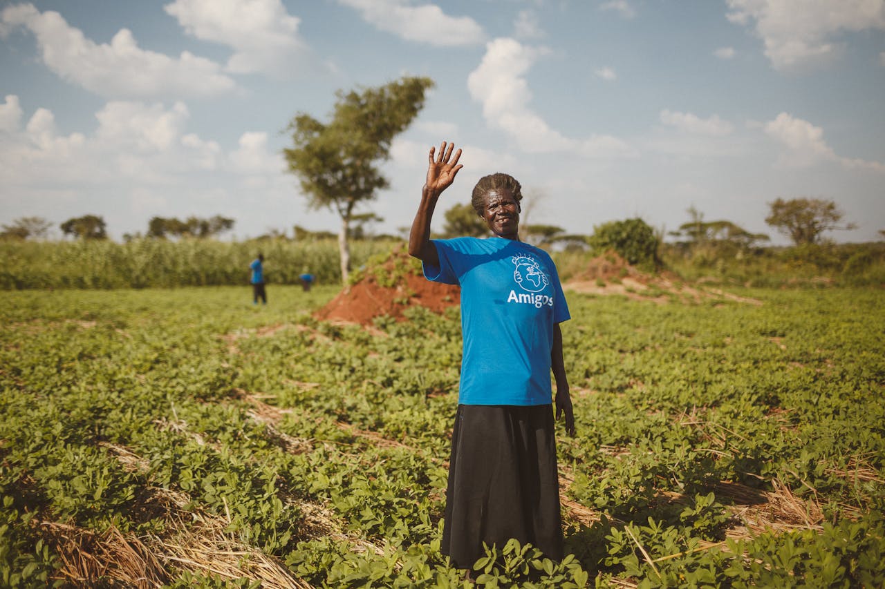 Elderly woman waves in a Ugandan field, showcasing rural agriculture and community spirit.