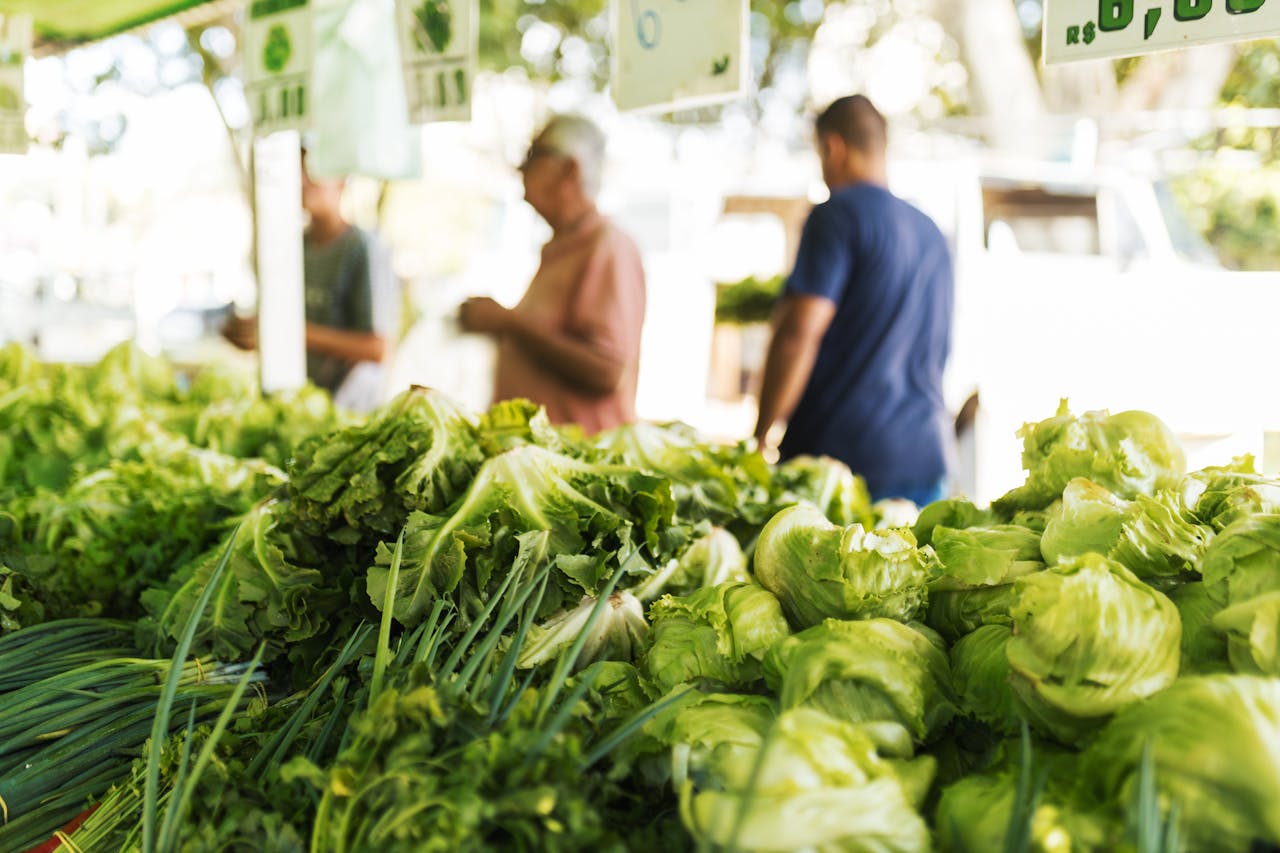 Vibrant display of green vegetables at an outdoor market with blurred shoppers in the background.
